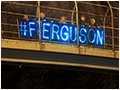 Protesters stand on a highway overpass with a lit up sign that reads "#FERGUSON".
