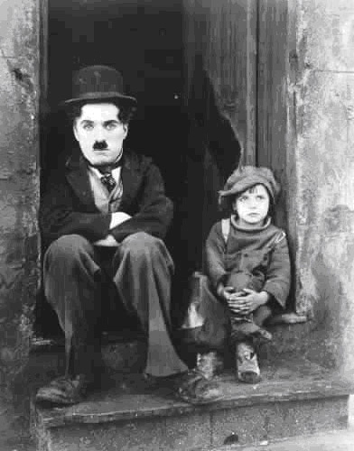 Film still of Charlie Chaplin 'Tramp' character sitting in a doorway beside a young child.