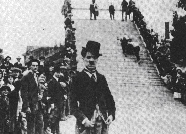 Film still of Charlie Chaplin, looking very similar to The Tramp character, with a ramp and crowd behind him.