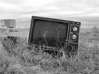 Black and white photo of a television abandoned in the dry prairie grass.