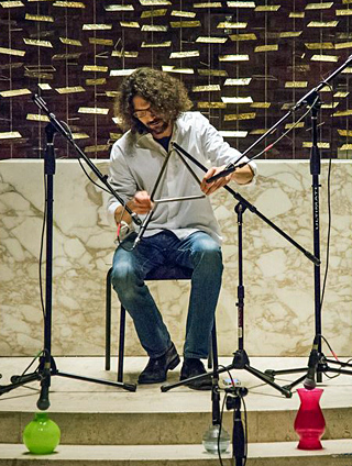 Photo of a seated man striking a metal triangle, with microphones.