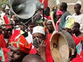 Photo of many people gathered in a street, with large loudspeakers, hand drums, and a hand holding up a tape recorder.