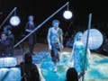 Puck pursues Peaseblossom across a clearing, surrounded by large paper lanterns on tall bamboo poles. An orchestra plays behind them.