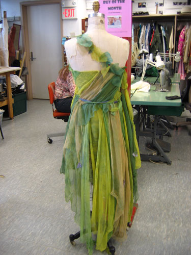 Seen from the back on a dress form, the dress has one strap coming across from the left shoulder to the bodice, which is made from silk strips in various shades of green, swept diagonally to the waist. The skirt falls raggedly past the knee, reaching the 