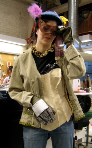 Over street clothes, a woman wears a corset mock-up, a jacket with painted Celtic knots, gauntlets with metal panels and wires, a gold-and-silver bead necklace, a purple-and-gold mask, and a blue hat with a yellow lily.