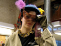 Over street clothes, a woman wears a corset mock-up, a jacket with painted Celtic knots, gauntlets with metal panels and wires, a gold-and-silver bead necklace, a purple-and-gold mask, and a blue hat with a yellow lily.
