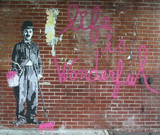 The surface of a red-brick building shows an illustration of a man with a small moustache, tight suit coat, and baggy pants, holding a paint can and roller. He stands next to the words "life is wonderful" painted in hot pink paint.
