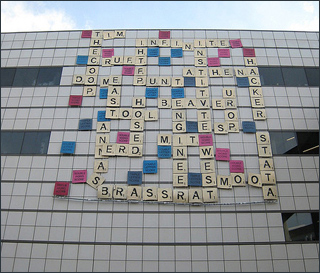 On the side of a building hang multi-colored squares, each with a letter or phrase on them, simulating a Scrabble game.