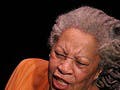 Toni Morrison holds up a book while she speaks at a lectern.