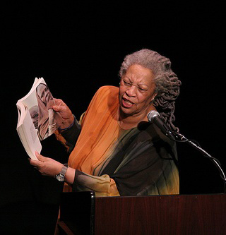Toni Morrison holds up a book while she speaks at a lectern.