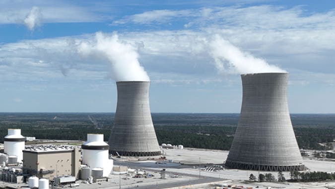 Two large concrete cooling towers at a power plant release white steam into a blue sky, with industrial buildings and storage tanks in the foreground.