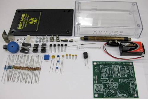 Photo of parts laid out on a table: plastic case, screws and fittings, Geiger Muller tube, 9V battery, discrete resistors and capacitors, printed circuit board.