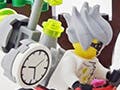 A LEGO® model of a young man on a bike, with a huge clock attached to his back, pointing a gun at an elegantly-dressed older gentleman.