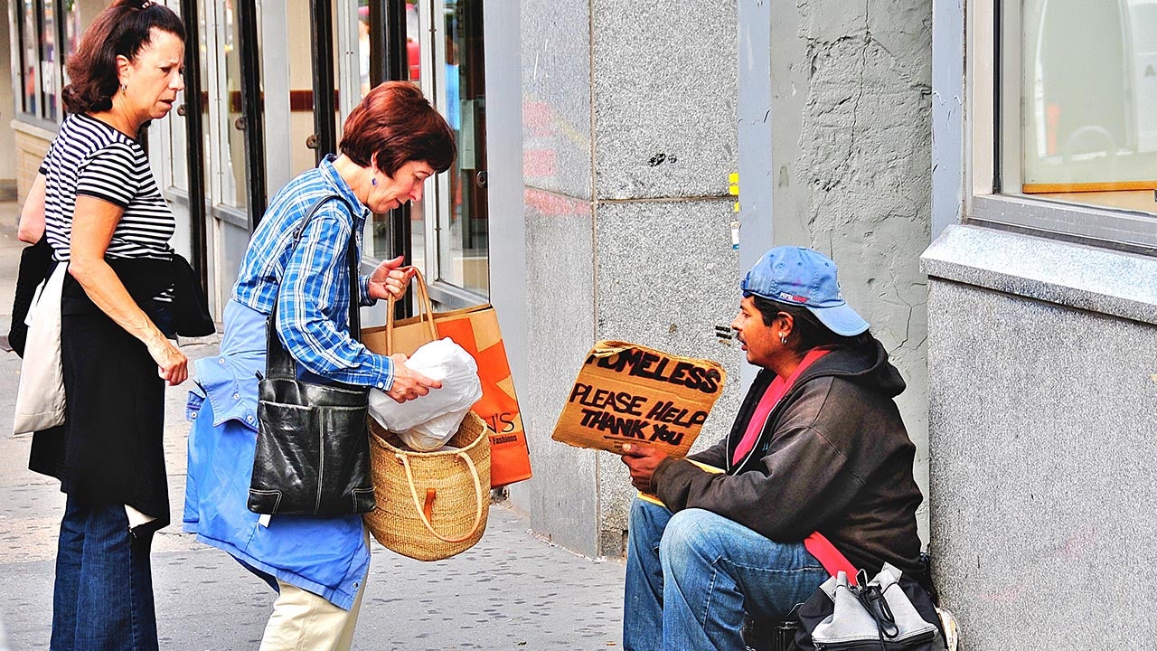 Seated on a duffel bag, a young man holds a sign with the words “Homeless. Please help. Thank you,” as a woman removes an object from one of her bags.