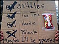 A young white man wearing a hoodie holds a sign over his head with the words  "Skittles," "Ice tea," and "Hoodie" listed next to boxes with check marks. Beneath them, next to a box with an x marked in it , are the words "Black. Maybe I'll be spared."