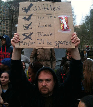 A young white man wearing a hoodie holds a sign over his head with the words  "Skittles," "Ice tea," and "Hoodie" listed next to boxes with check marks. Beneath them, next to a box with an x marked in it , are the words "Black. Maybe I'll be spared."