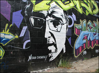 A mural on a brick wall depicts a half-lit bespectacled man's face, surrounded by  multi-colored script.
