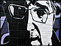 A mural on a brick wall depicts a half-lit bespectacled man's face, surrounded by  multi-colored script.