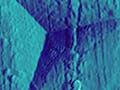 A sharp probe has left a triangular indentation in a surface, shown using scanning electron microscopy.