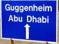 A photograph that features a large blue sign in the foreground that reads "Guggenheim Abu Dhabi." A construction site with cranes can be seen in the background.