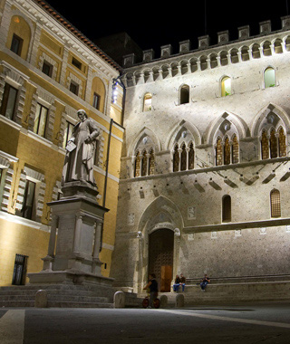 The Piazza Salimbeni in Sienna, Italy at night, showing the facade of the Banca Monte dei Paschi di Siena,Tito Sarrocchi’s statue of Sallustio Bandini, and several people, including a child on a bicycle.