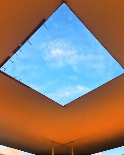 A photo of a ceiling with a square opening revealing blue skies and clouds above.