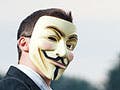 A man in a suit wears a Guy Fawkes mask on his face while standing next to a computer.