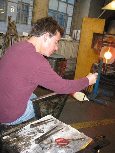 Photo of the glassblower shaping the end of the vase with a pair of tongs.