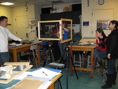 Photo of a string running from the lute [left] through the picture frame which is crosswise to the photograph's view and to the wall; students look through the picture frame. 