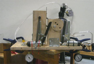 A hand-powered vacuum pump features a wooden flywheel that connects to two commercial vacuum pump units, mounted on a wooden frame.