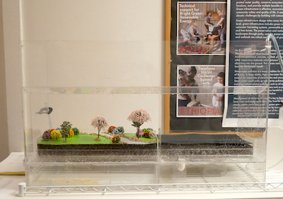 Diorama with falling rain; park setting on the left and asphalt on the right. 