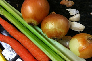 A photograph of garlic, onions, celery, and carrots lying on a counter.