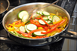 A frying pan containing peppers and zucchini sits on the burner of a stove.