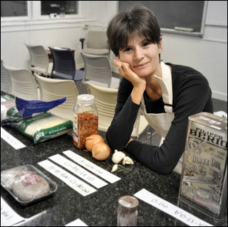 A woman poses in a kitchen with various items like olive oil, garlic and onions.