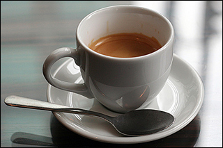 A tiny white cup of espresso sits on a white saucer next to a spoon.