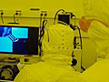 People in cleanroom suits view magnified wafer pattern on screen.