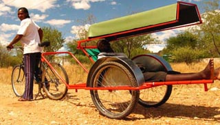 Photo of a two-wheeled ambulance cart attached to rear of bicycle, carrying a reclining patient.