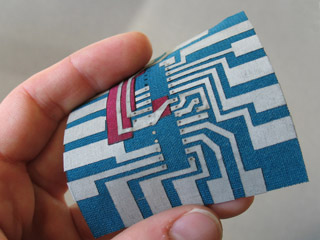Photo of a hand holding a small fabric square with a "printed circuit" pattern of connectors on it.