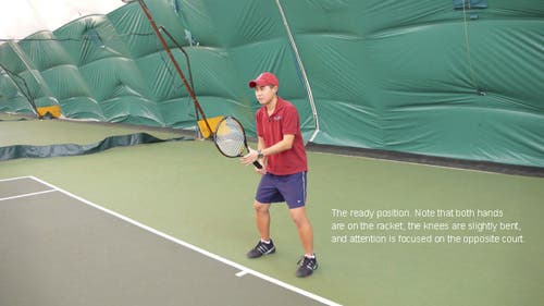 The player holds the racket in the right hand, with the head aligned straight up and down, tilted forward toward the net, and the left hand lightly steadying it at the neck.