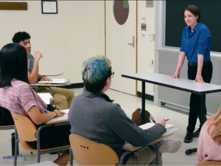 AN instructor in a classroom stands facing four college-aged students