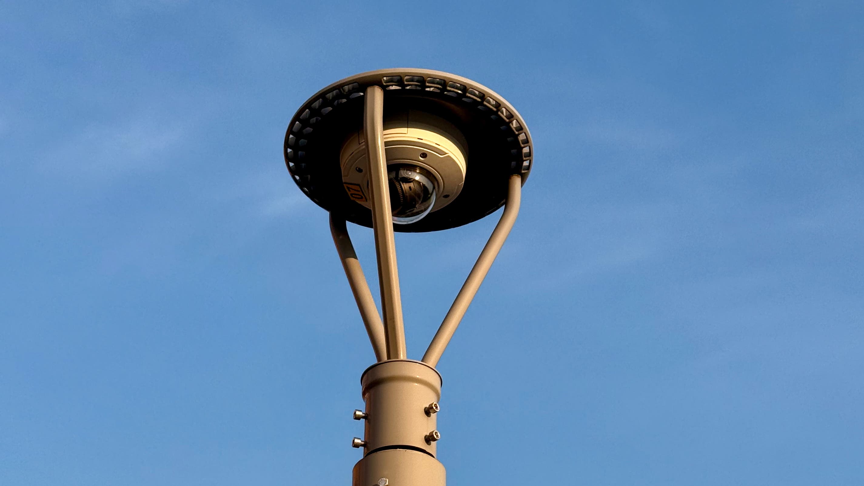 A surveillance camera atop a metal pole, against a blank blue sky.