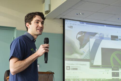 A man speaks into a microphone. There is a screen playing a YouTube video in the background.