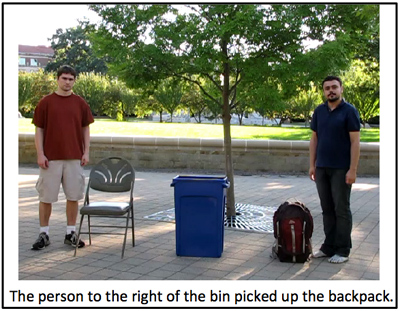 Still photo of a tall blue bin in center, with a man to the left standing with a folding chair and a man to the right with a backpack.