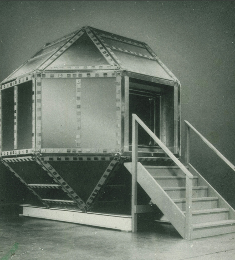 A black and white photo of a shielded hexagonal metal chamber atop two metal girders with a man sitting inside.