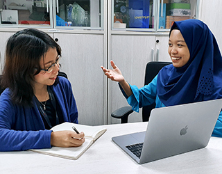 Two women sit together at a table talking to each other. One woman is taking notes with a pen and notebook. The other woman sits in front of an open laptop.  