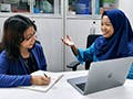 Two women sit together at a table talking to each other. One woman is taking notes with a pen and notebook. The other woman sits in front of an open laptop.  