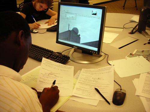 A student sits a table with a computer screen showing MIT's Doppler ball, while he writes on a piece of paper. 