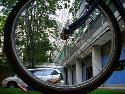 A trick photo that shows a bike tire in the foreground and a person lying in the grass in the background. It looks like the person is getting run over by the bike. 