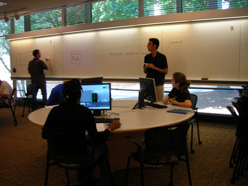 A photo of a classroom with a teacher at the front, a student at the whiteboard, and two students at a round table. 