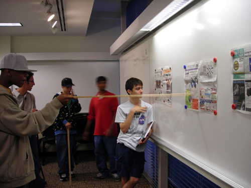 Newspapers are taped on a whiteboard. Students try reading them from different distances from the whiteboard. 